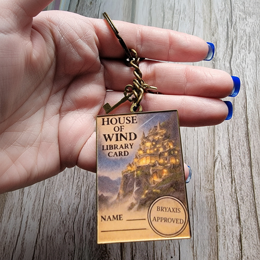 Hand holding a keychain with a 'House of Wind Library Card' design on a wooden surface