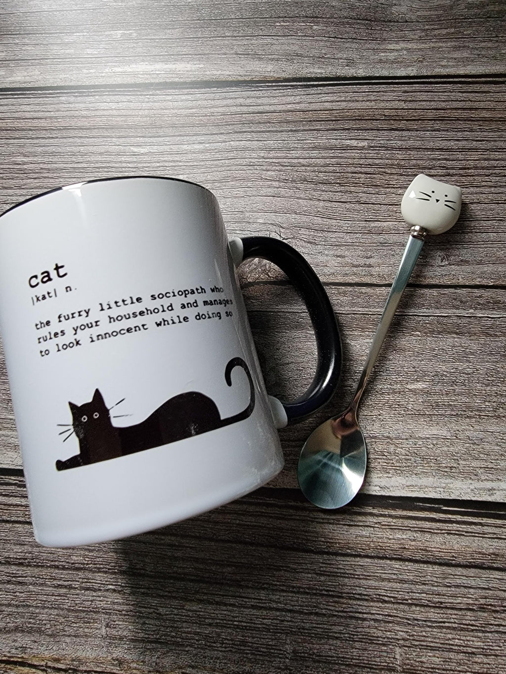 White mug with black cat illustration and text on a wooden surface with a spoon and decorative item.