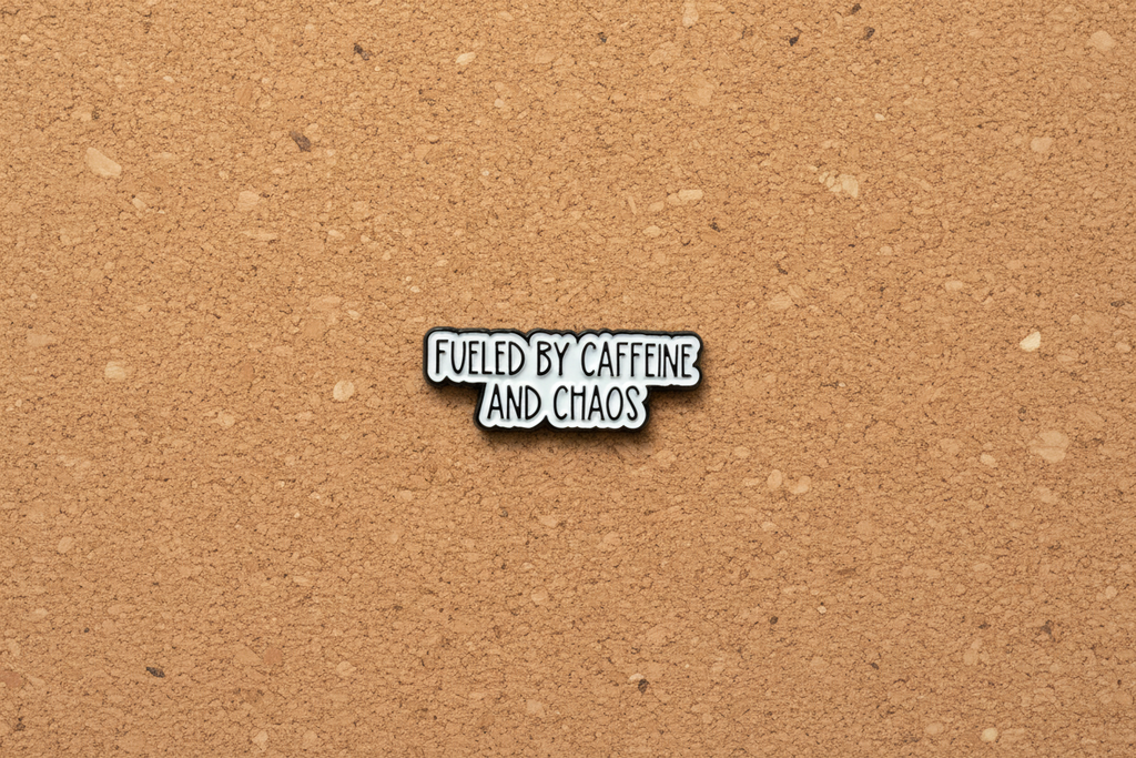Metallic badge with text 'Fueled by caffeine and chaos' on a cork background