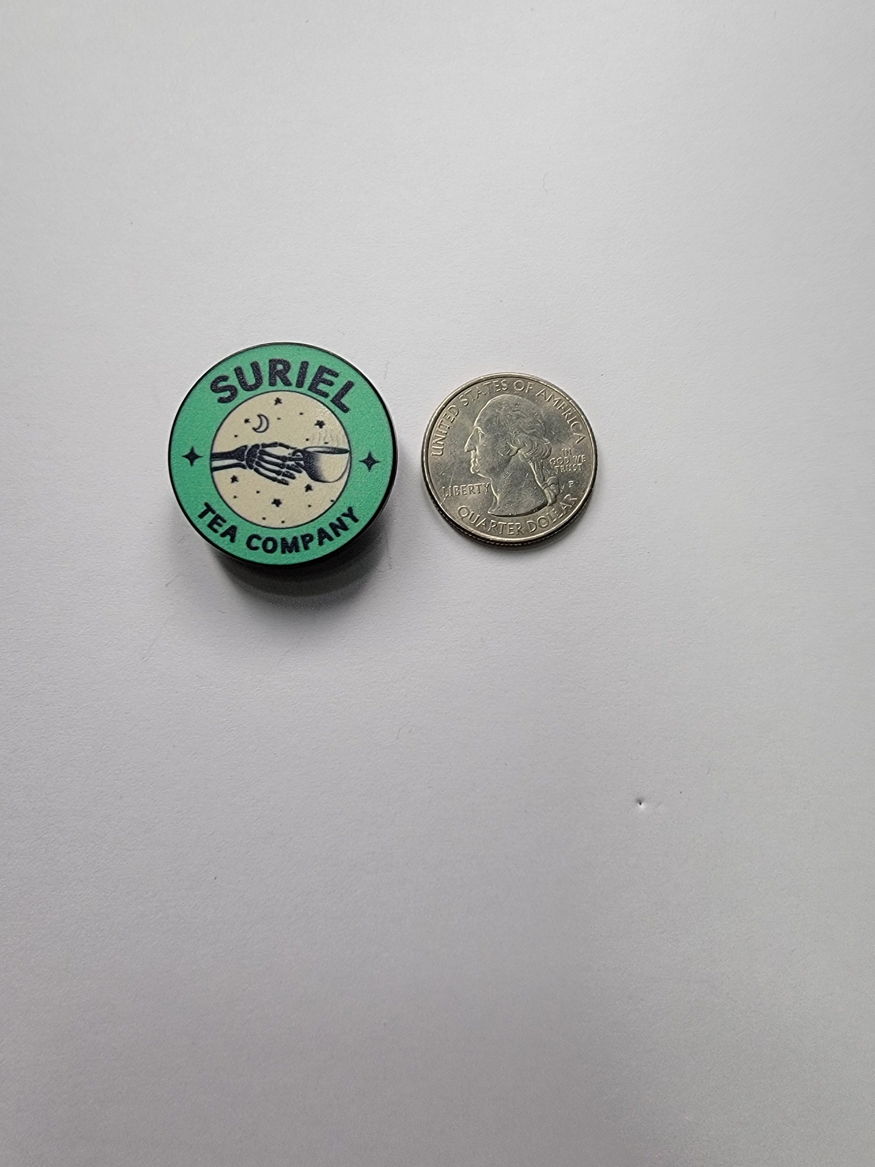 Small round green and black badge with 'Suriel' text next to a coin on a gray background