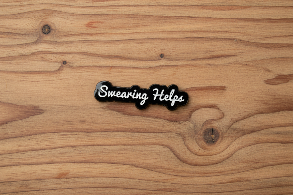 Black and white enamel pin that say s "swearing helps" on a wood background