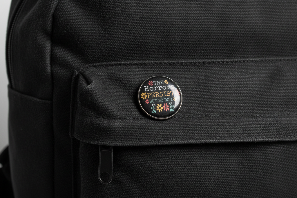 Round black pin with text and floral designs on a light gray bookbag background