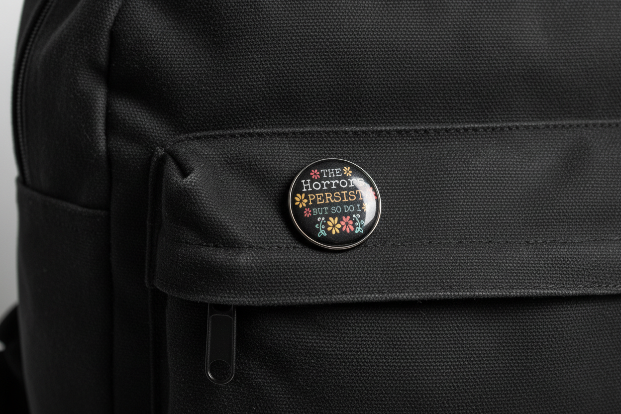 Round black pin with text and floral designs on a light gray bookbag background