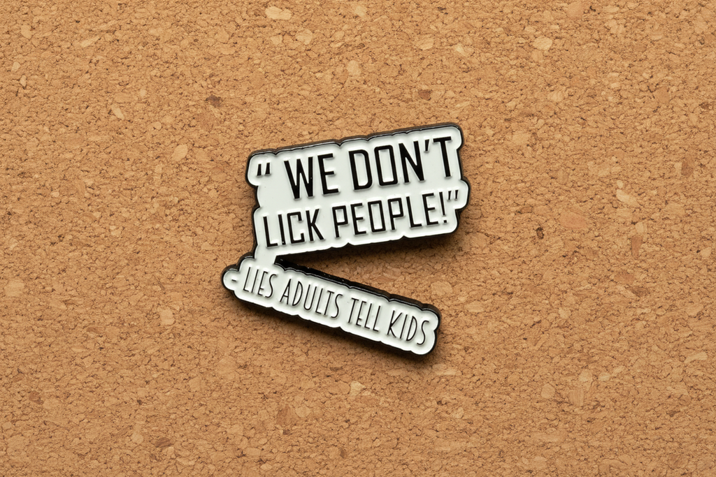 a white enamel pin with black text that says we don't lick people lies adults tell kids on a cork background