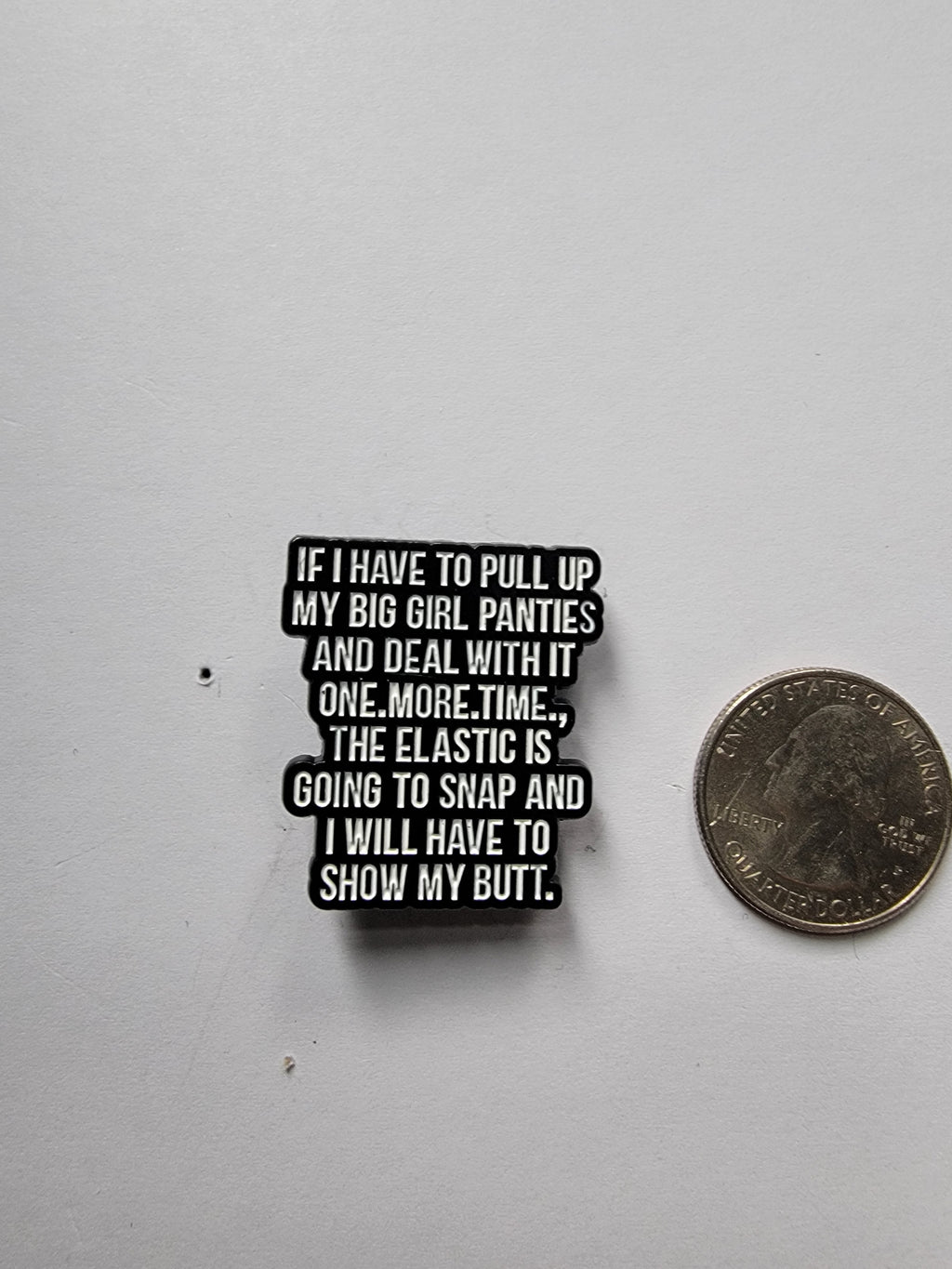 a black enamel pin with white text next to a quarter on a white background