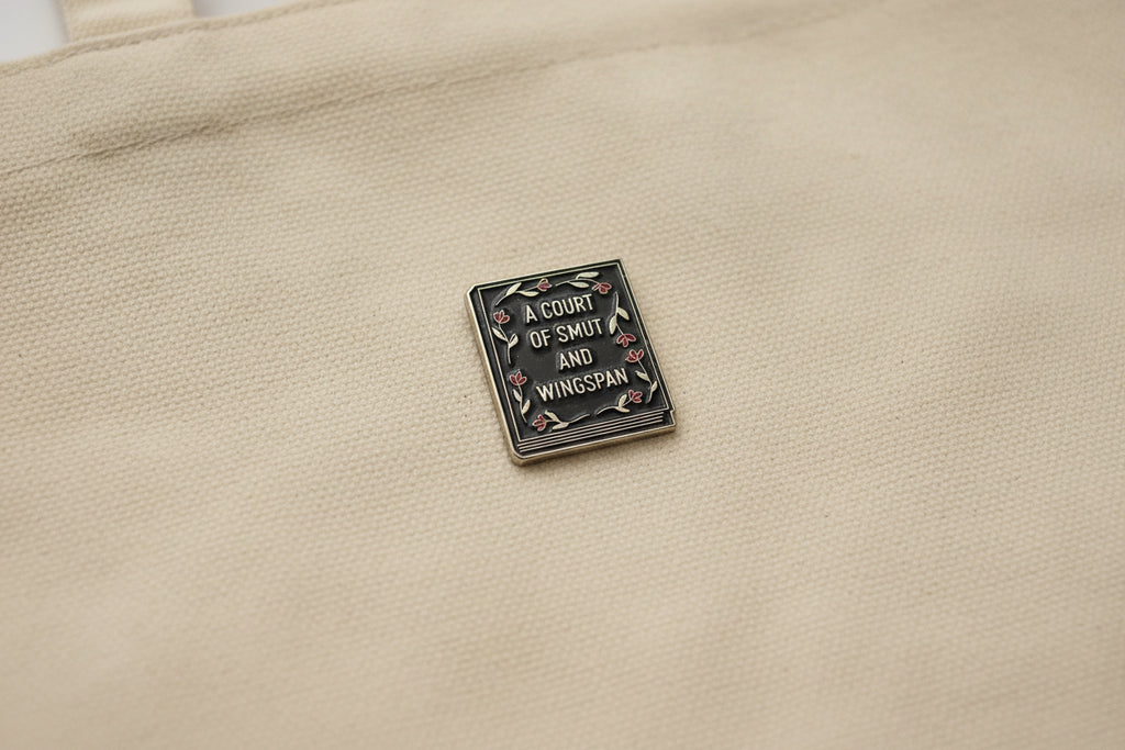 Enamel pin with 'A Court of Smut and Wingspan' text on a beige fabric background