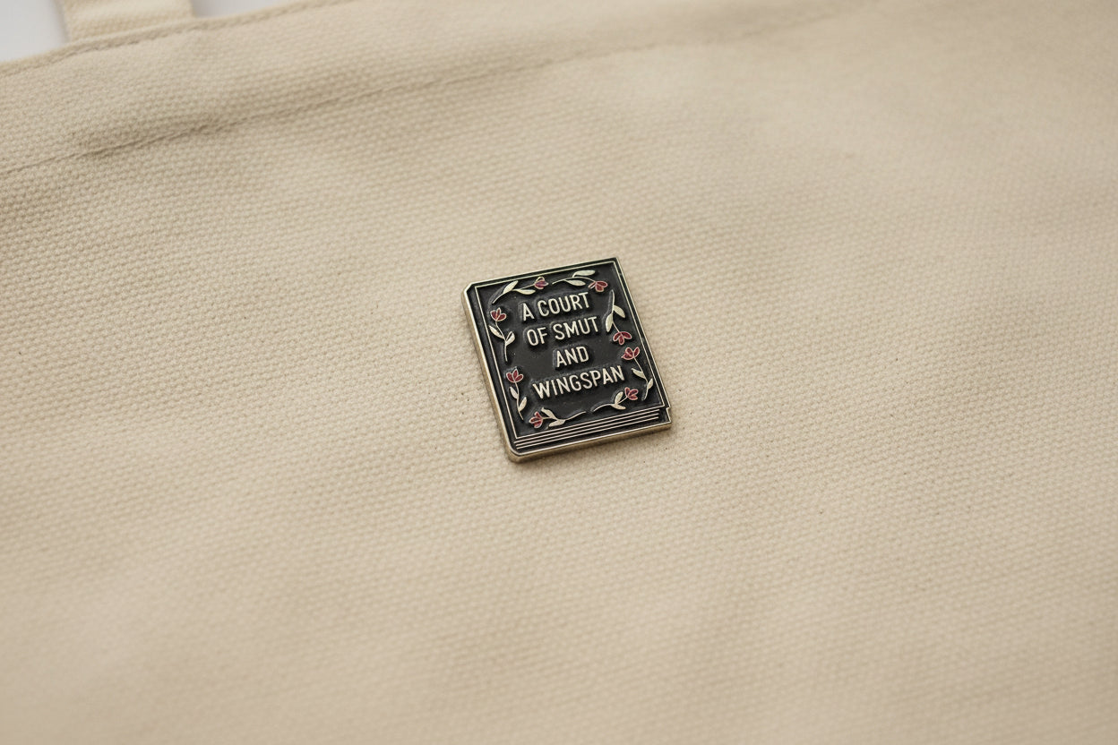 Enamel pin with 'A Court of Smut and Wingspan' text on a beige fabric background