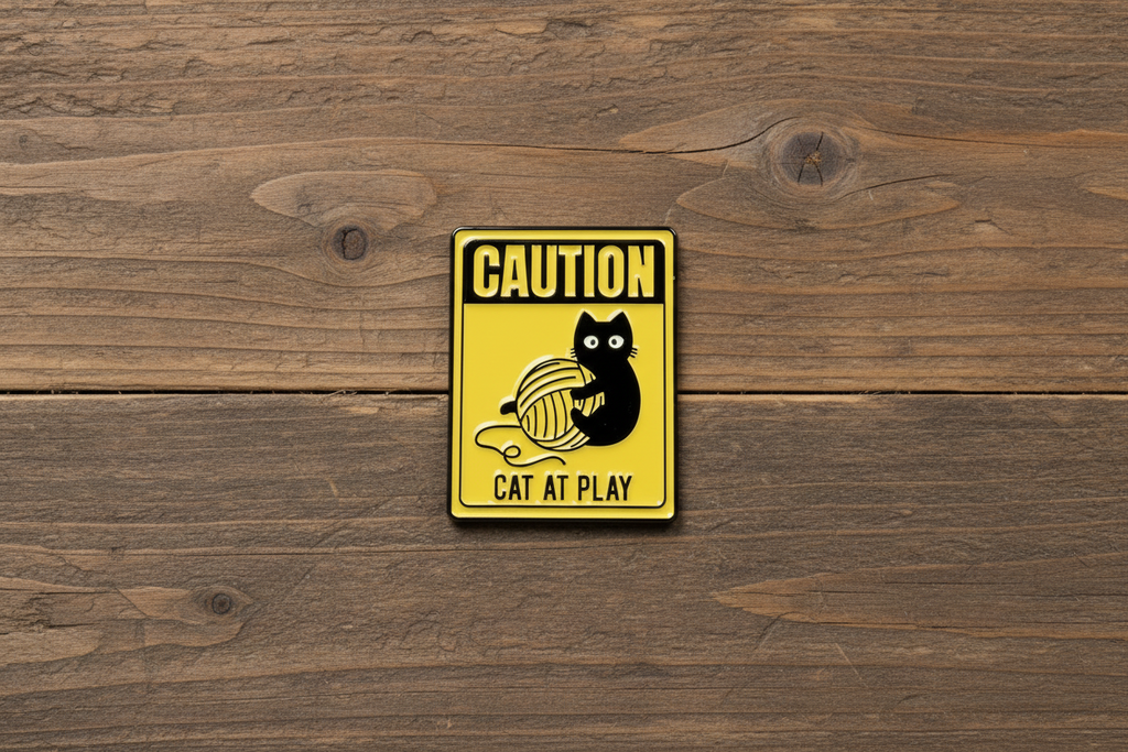 a yellow enamel pin with a cat playing and text on a wood background