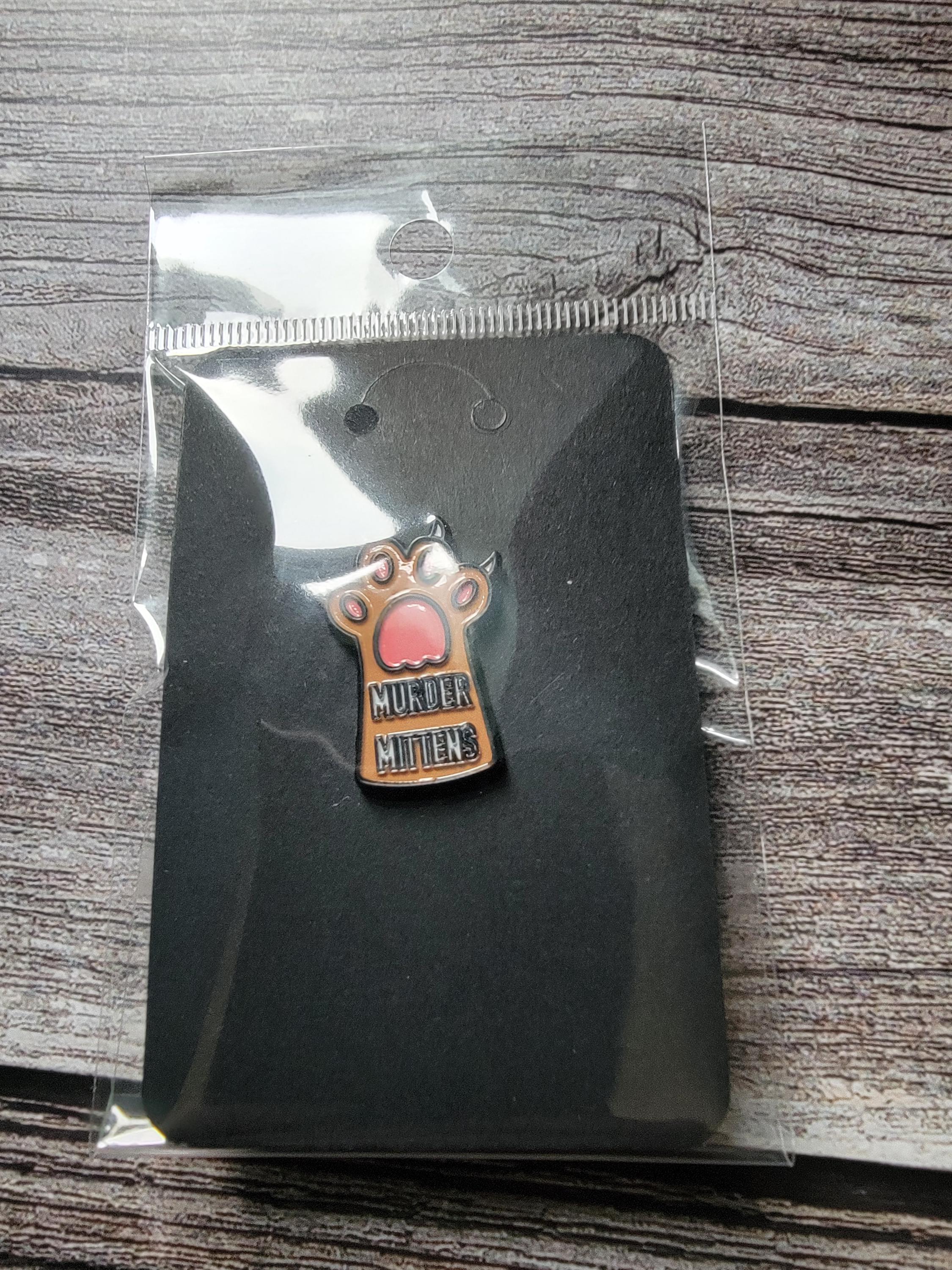 Pin with paw design and 'Murder mittens' text on a black card, in clear packaging.