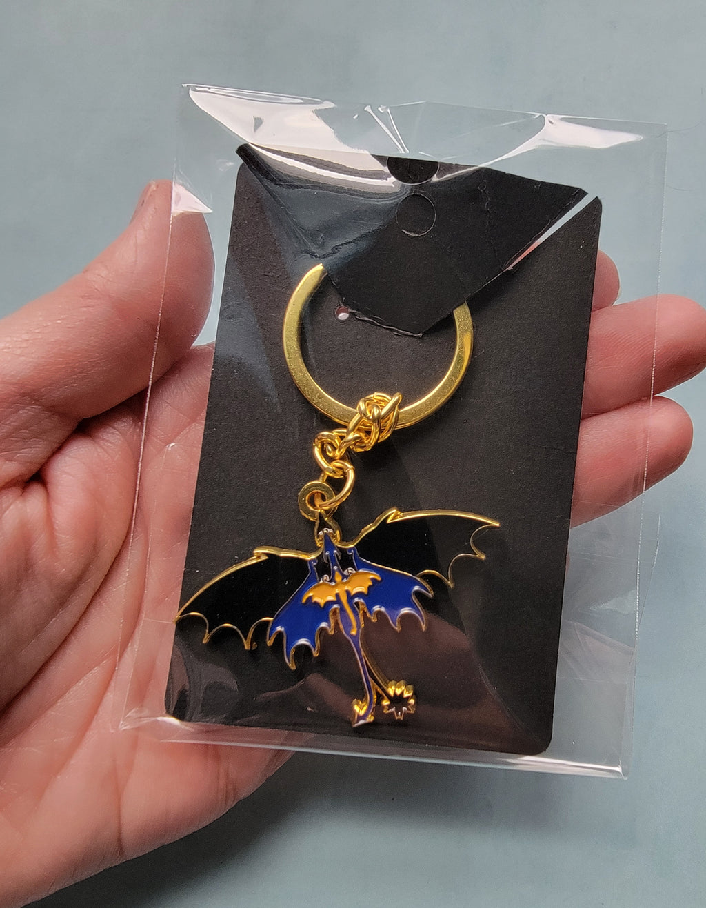 Gold and blue dragon-shaped keychain in clear packaging held by a hand against a gray background