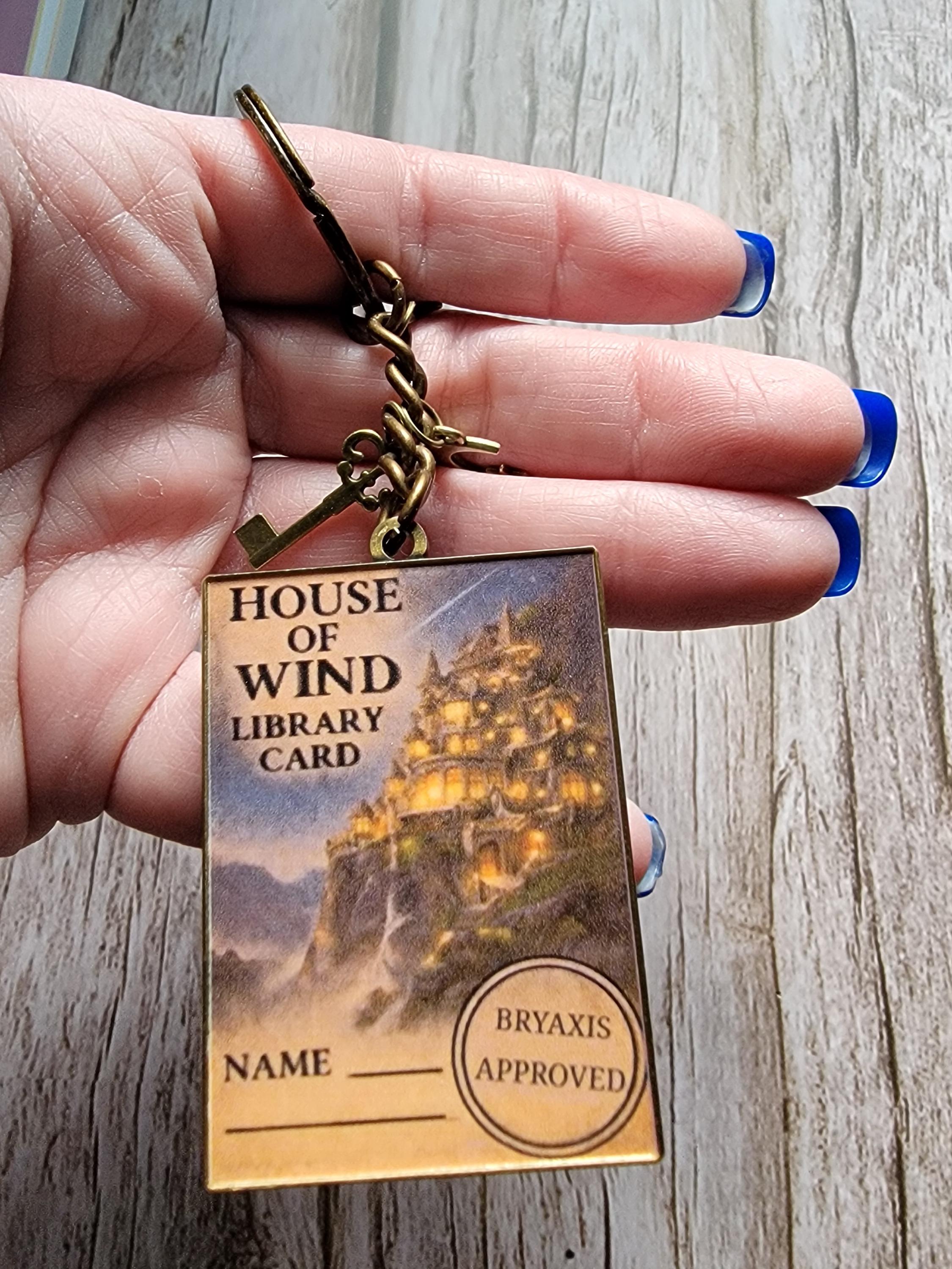 Hand holding a keychain with a 'House of Wind Library Card' design on a wooden surface