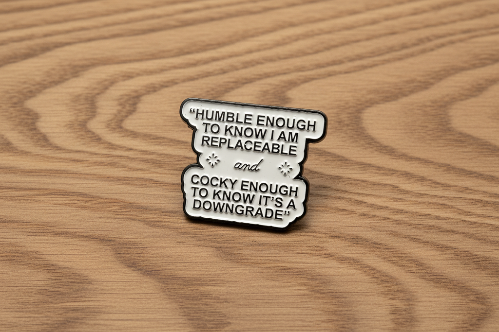 white enamel pin with black text on a wood background
