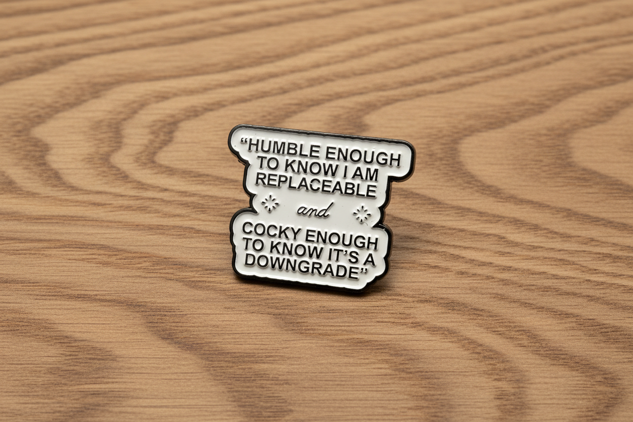 white enamel pin with black text on a wood background

