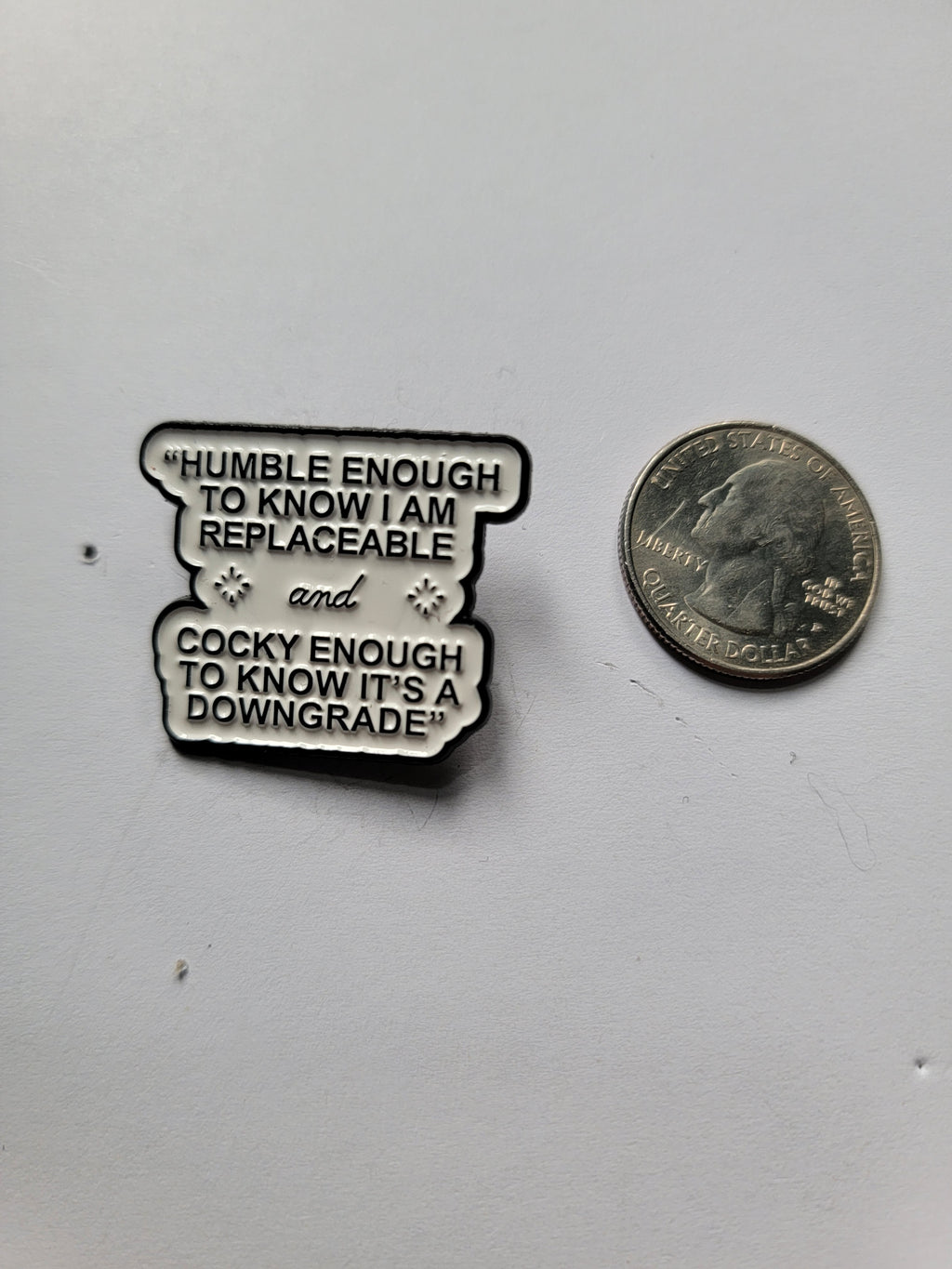 white enamel pin with black text next to a quarter on a white background
