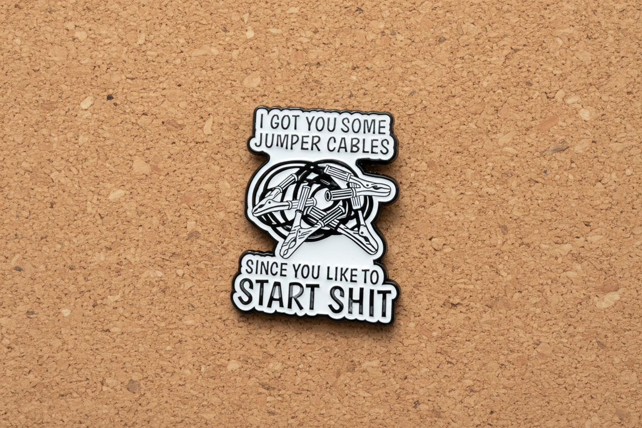 white enamel pin with black text and jumper cables on it on a cork background