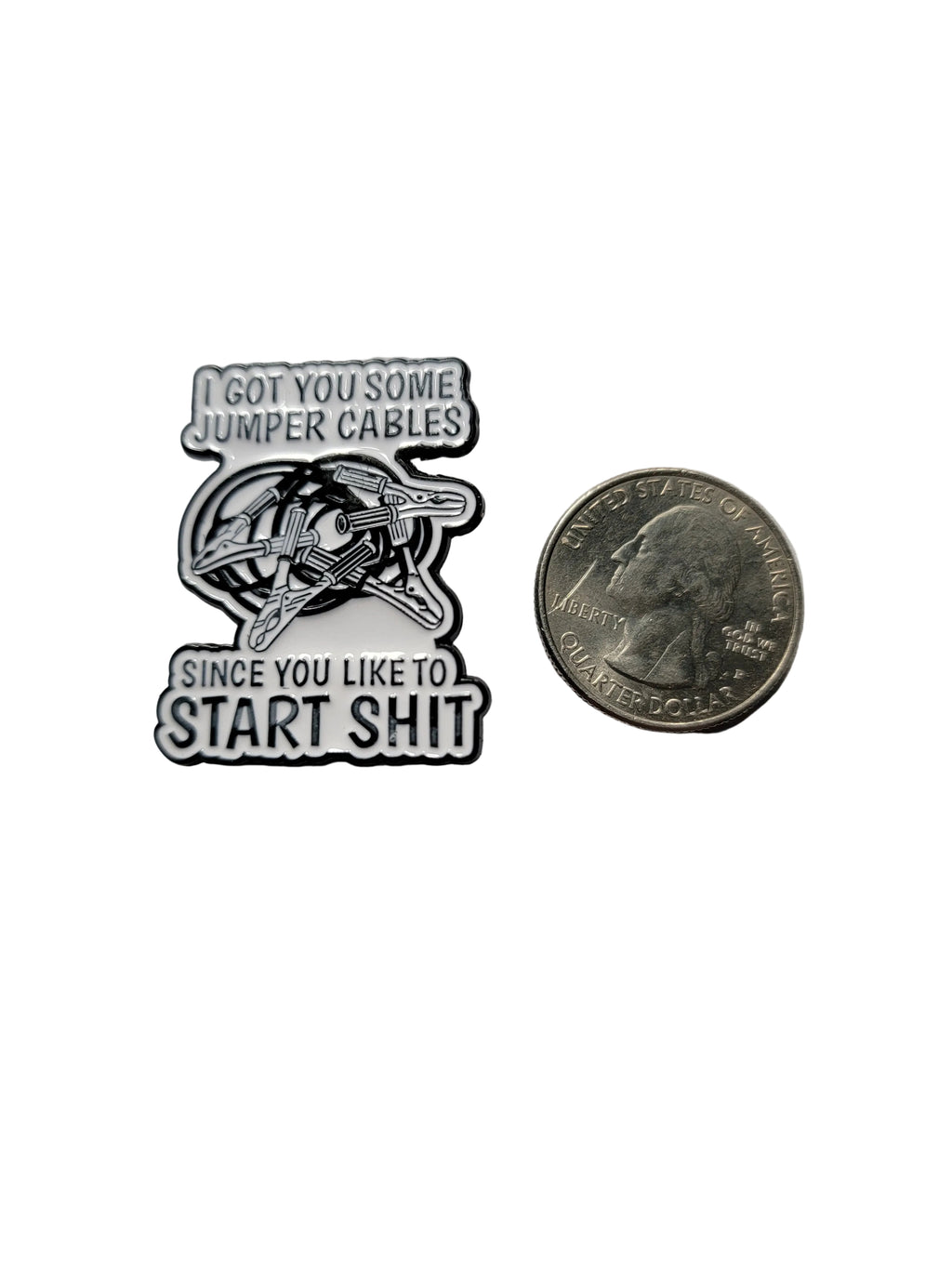 a white enamel pin with black text and jumper cables next to a quarter on a white background