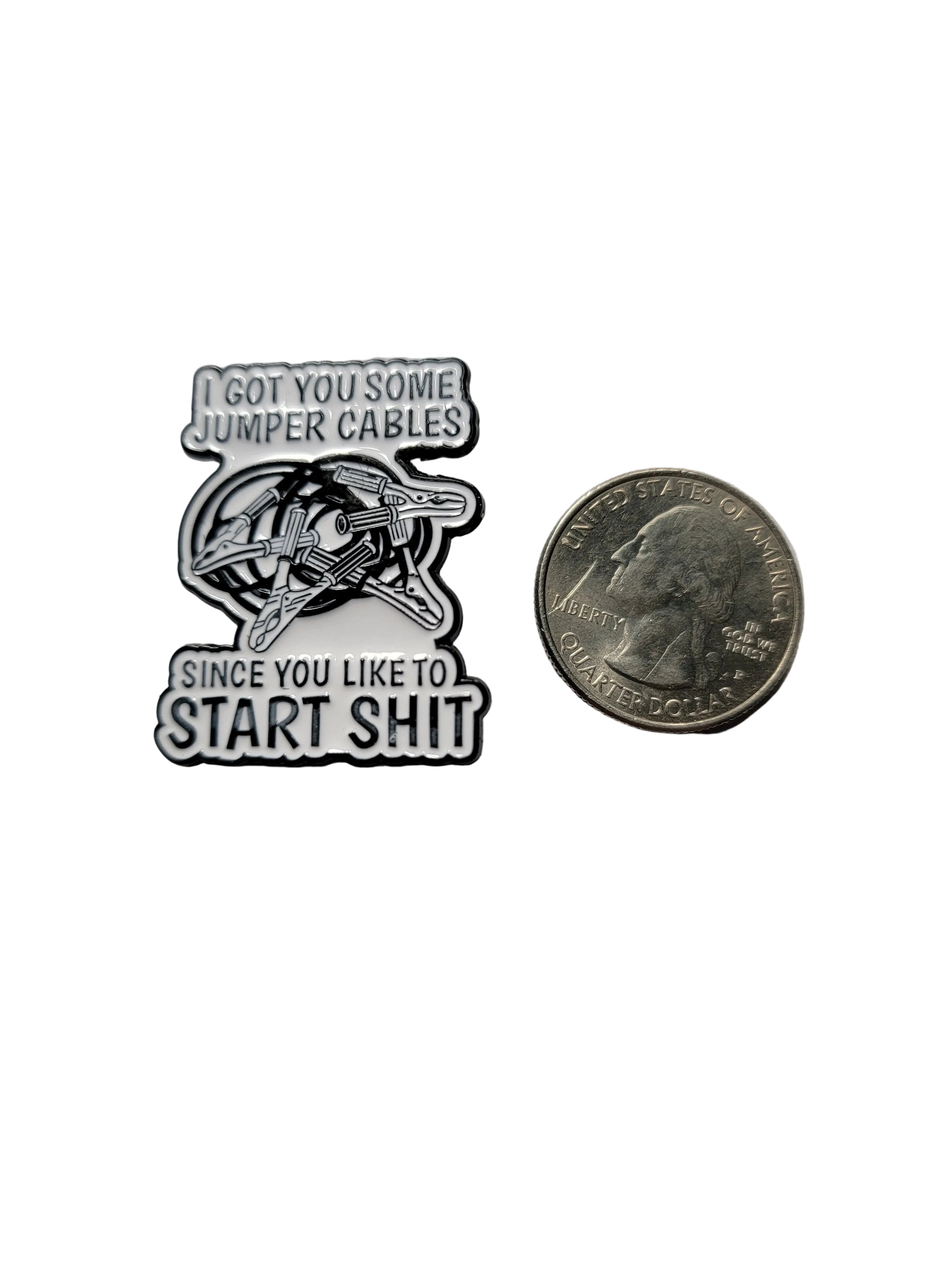 a white enamel pin with black text and jumper cables next to a quarter on a white background