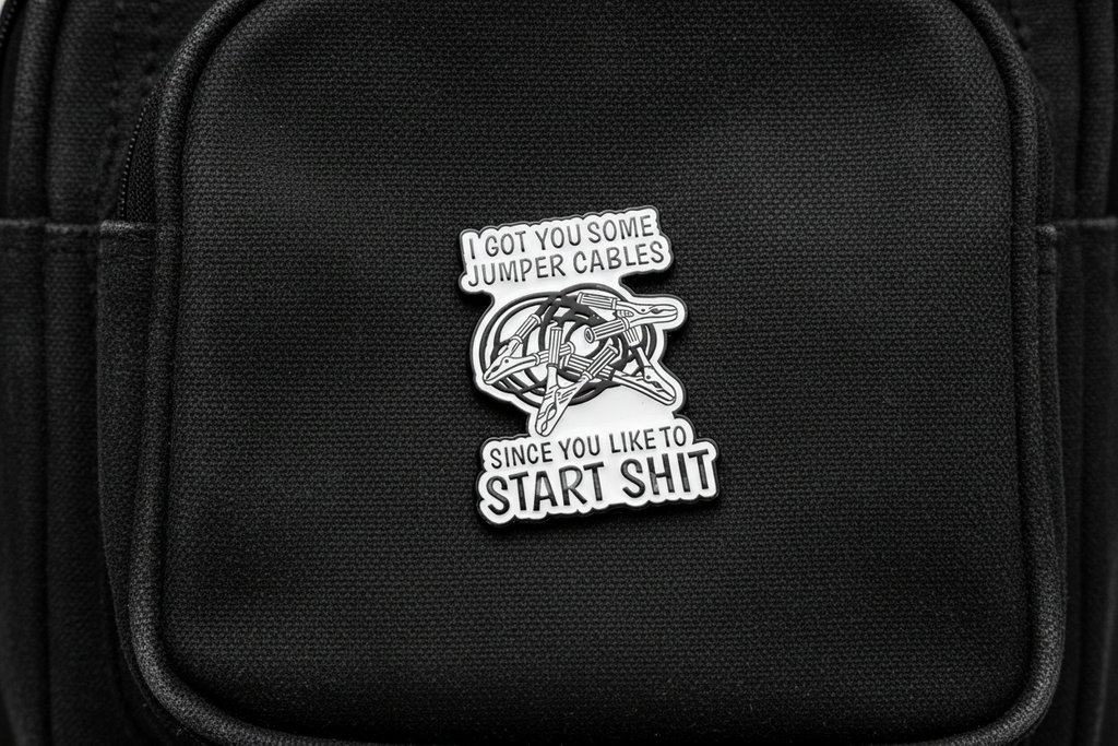 white enamel pin with black text and jumper cables on it on a black bookbag background