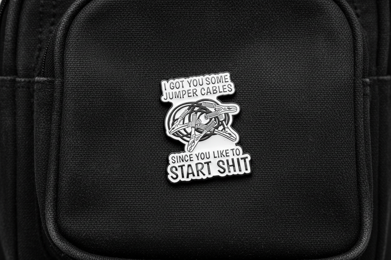 white enamel pin with black text and jumper cables on it on a black bookbag background