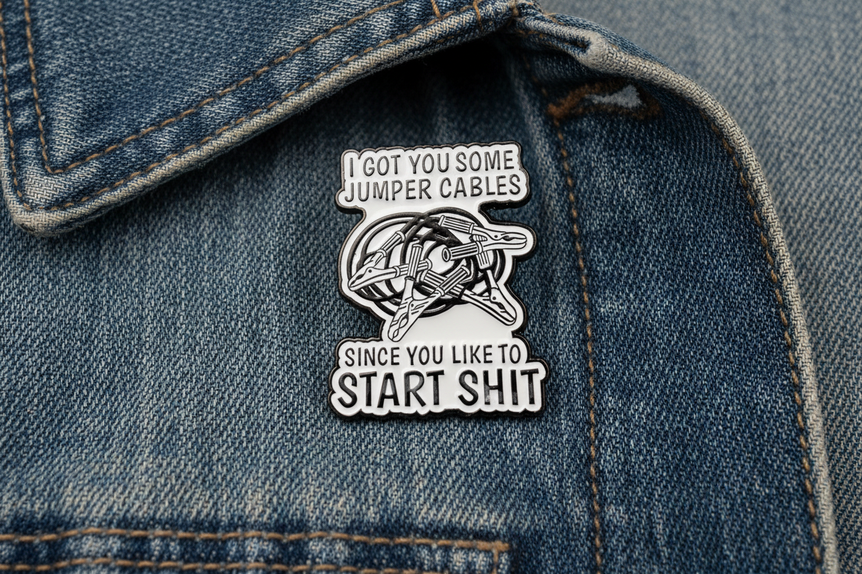 white enamel pin with black text and jumper cables on it on a denim background