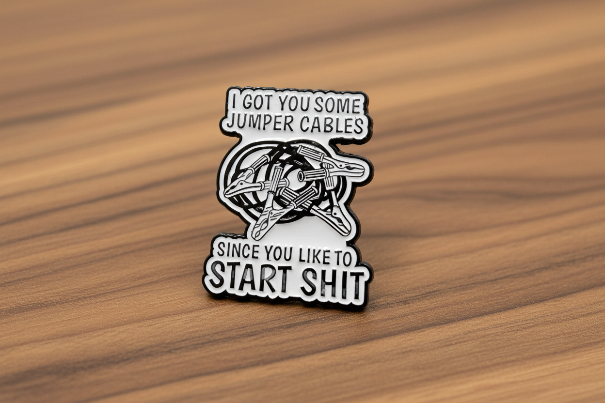 white enamel pin with black text and jumper cables on it on a wood background
