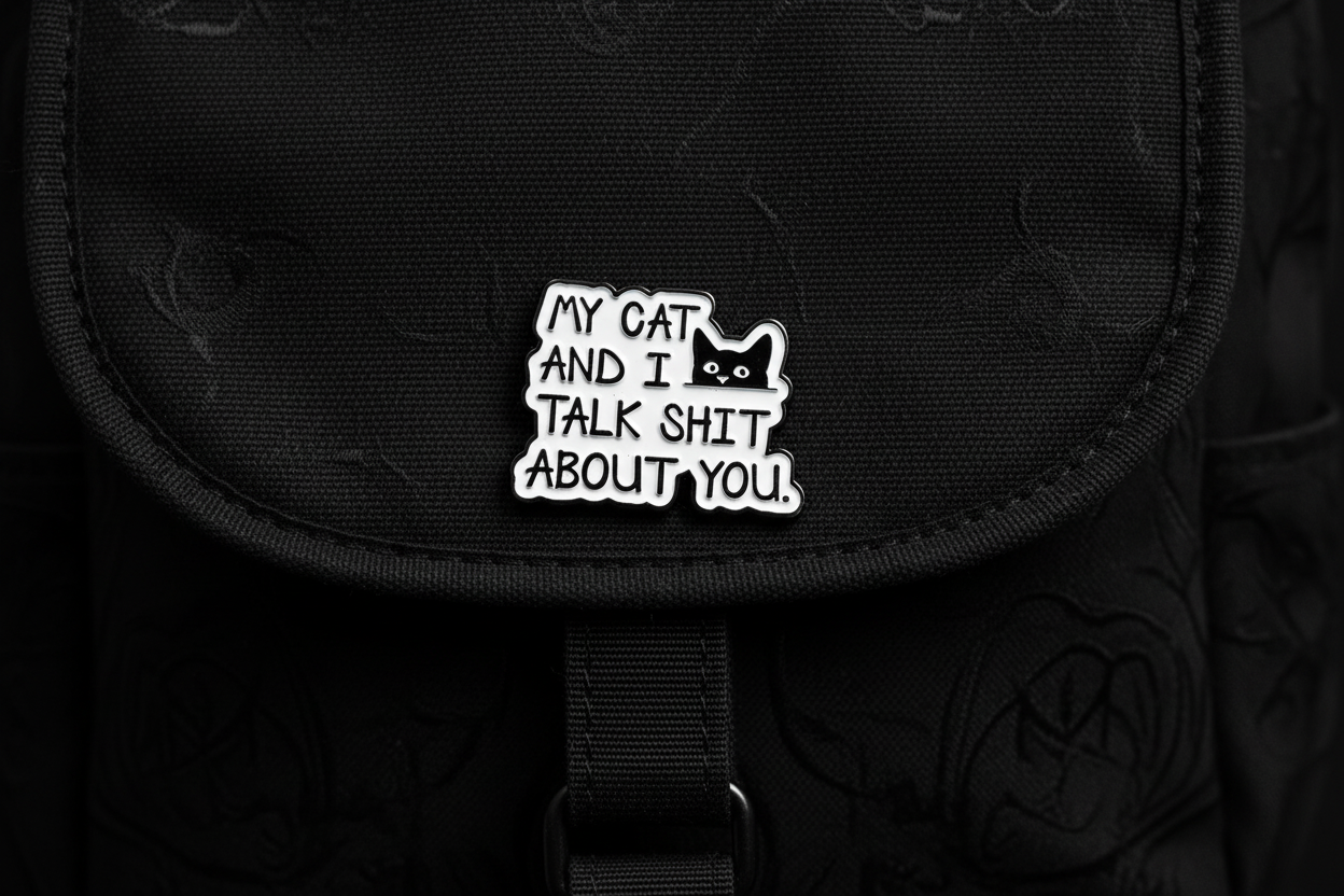 Metallic badge with text 'My cat and I talk shit about you' on a black bookbag background