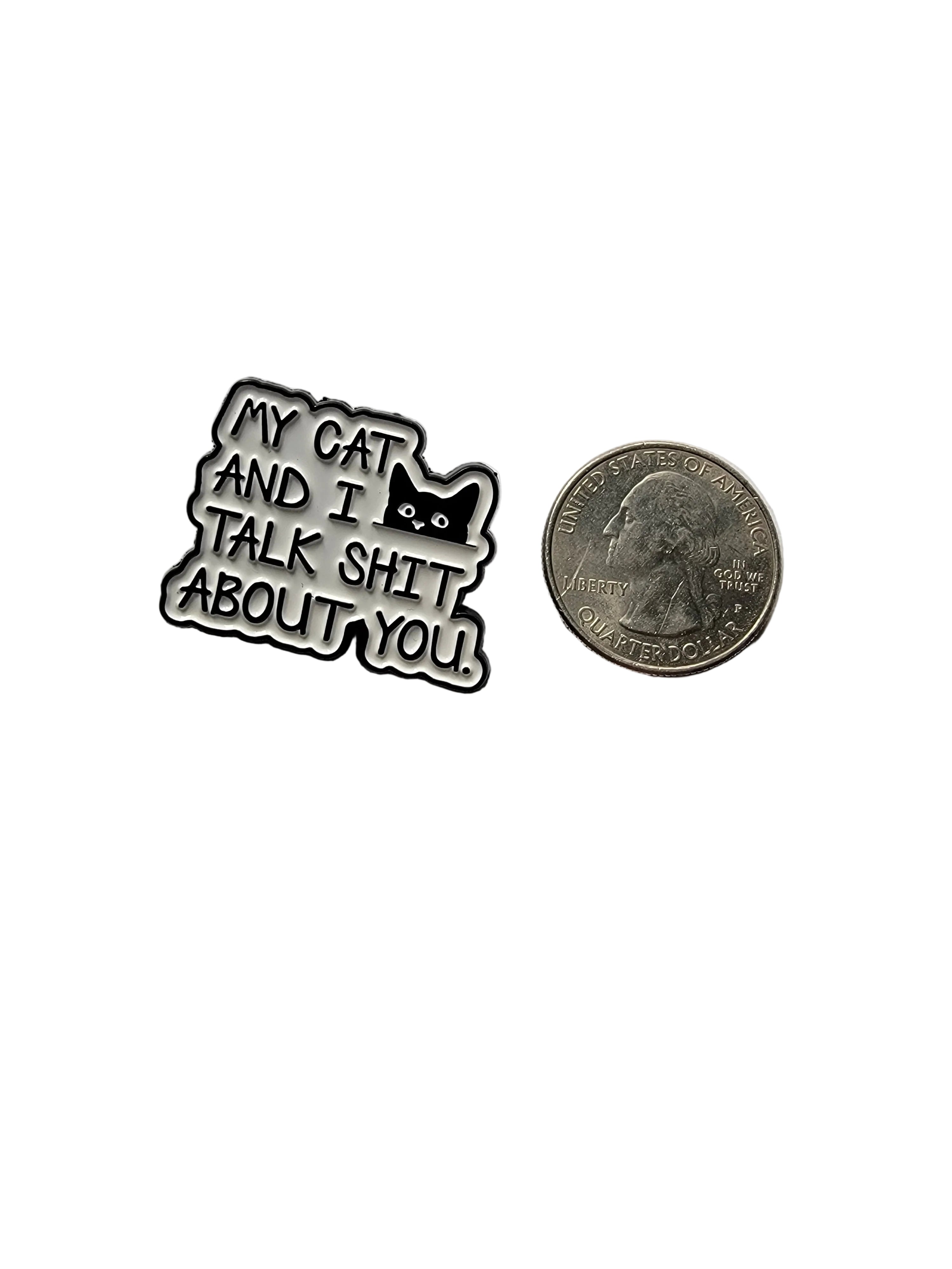 a white and black enamel pin with text and a cat next to a quarter for scale on a white background 