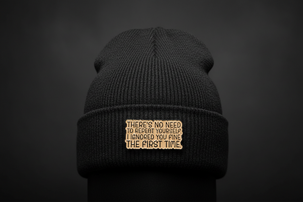 a beige enamel pin with black text on a black beanie background