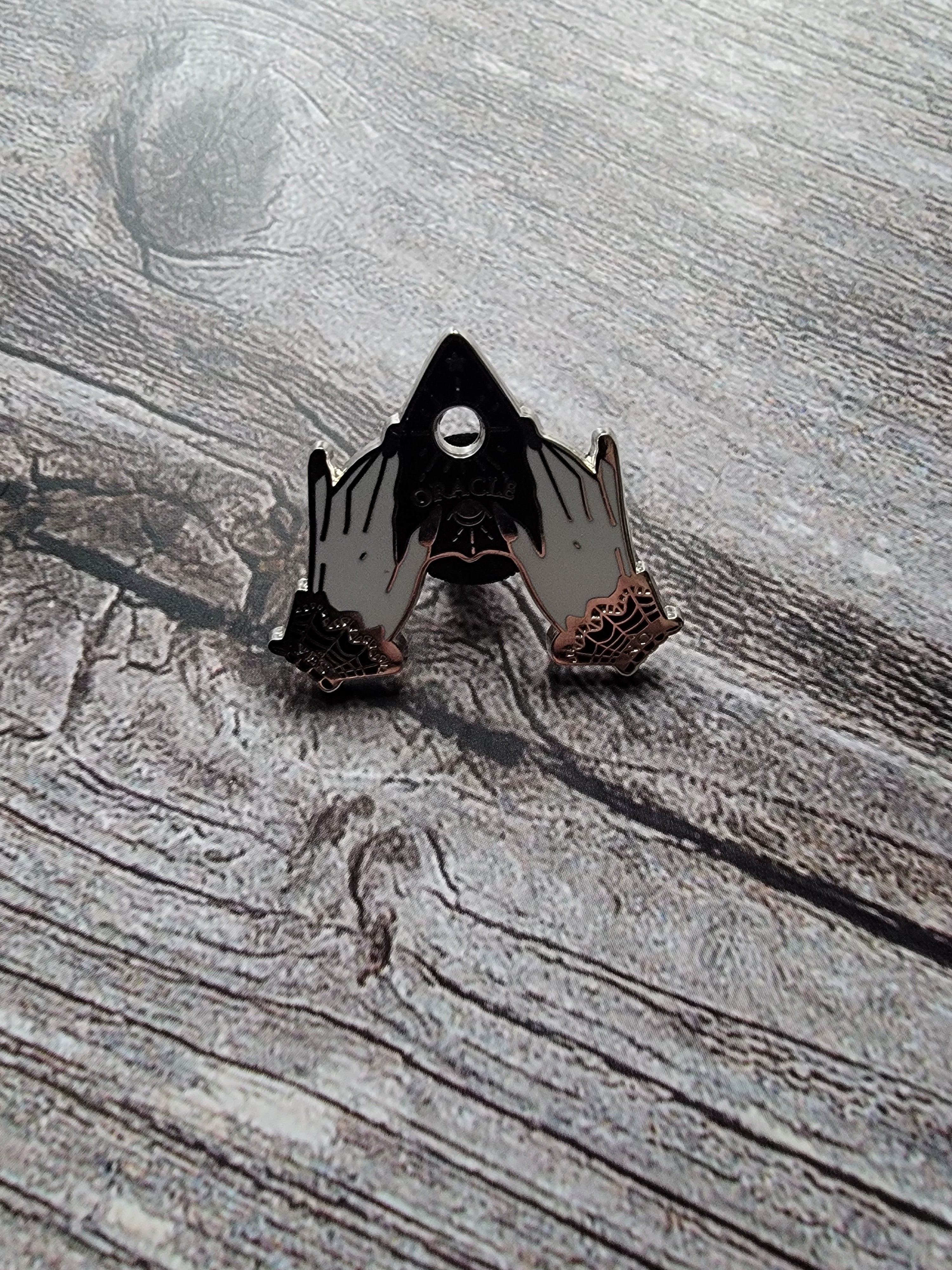 planchette enamel pin on wood background gothic enamel pin with hands holding a planchette 