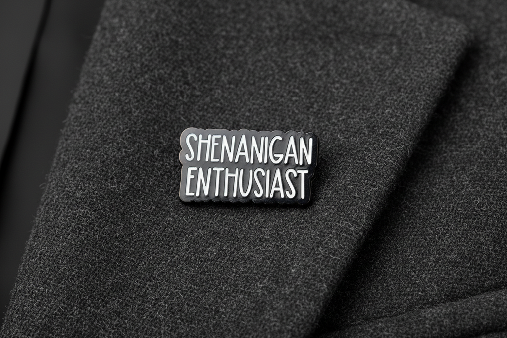 Black and white enamel pin with 'SHENANIGAN ENTHUSIAST' text on a gray fabric background