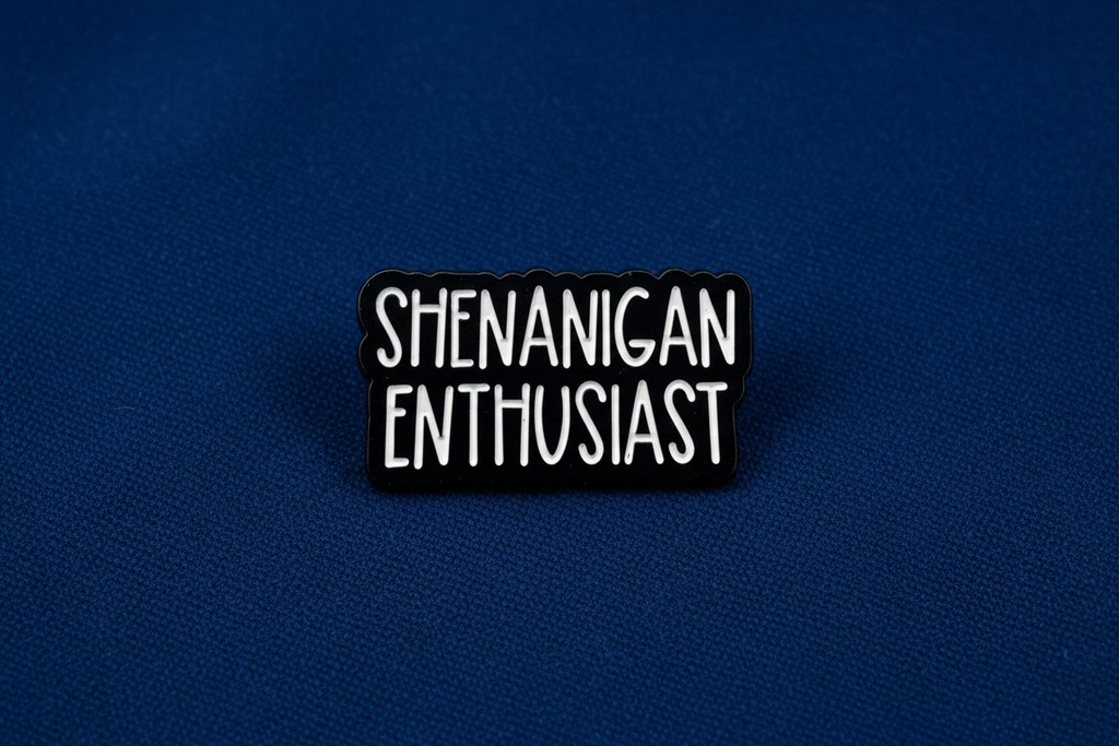 Black and white enamel pin with 'SHENANIGAN ENTHUSIAST' text on a blue fabric background
