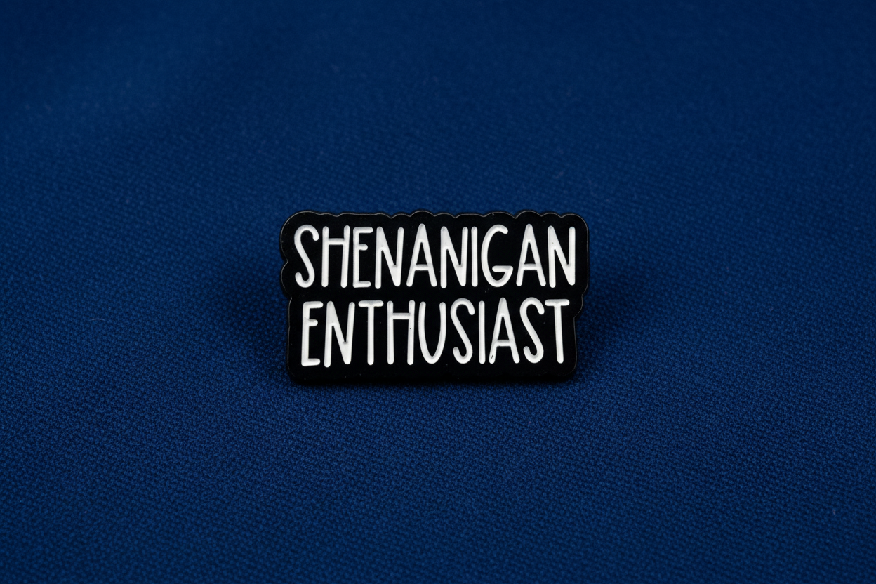 Black and white enamel pin with 'SHENANIGAN ENTHUSIAST' text on a blue fabric background
