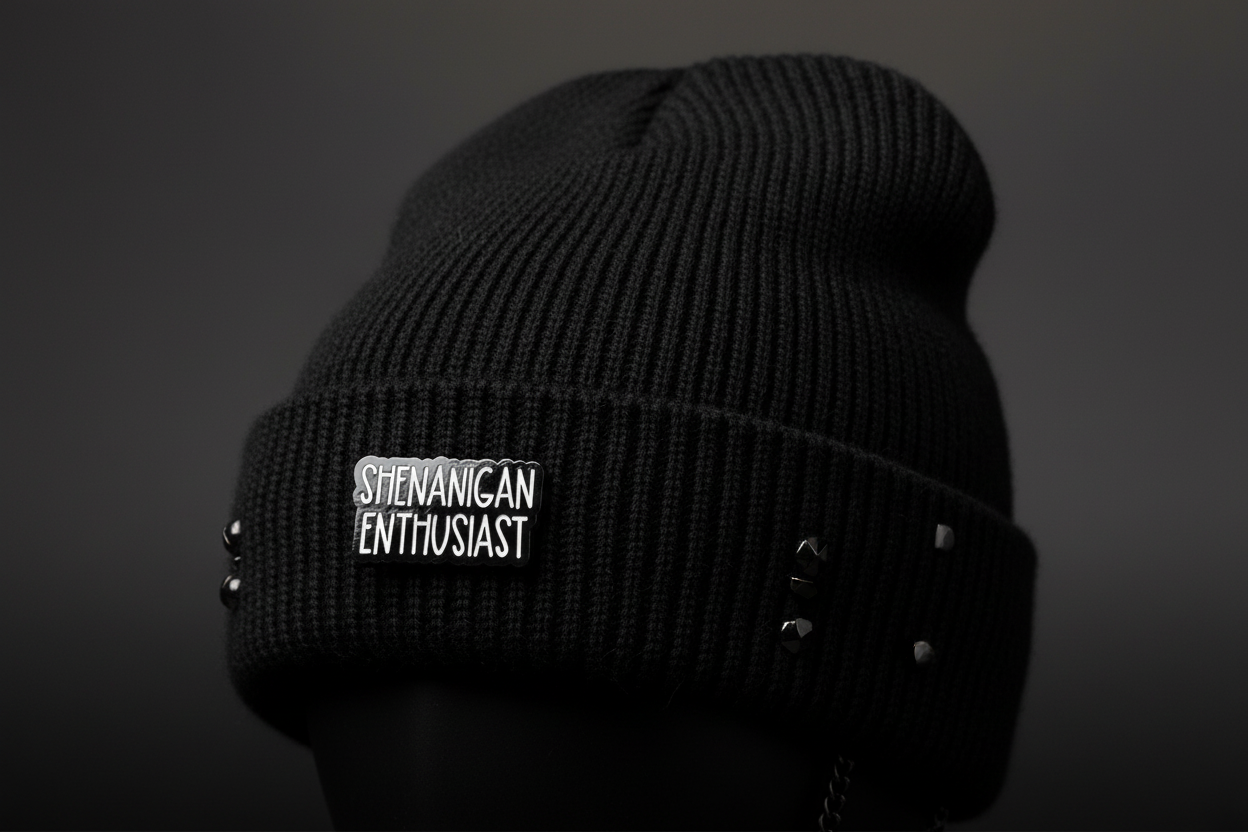 Black and white enamel pin with 'SHENANIGAN ENTHUSIAST' text on a black beanie background