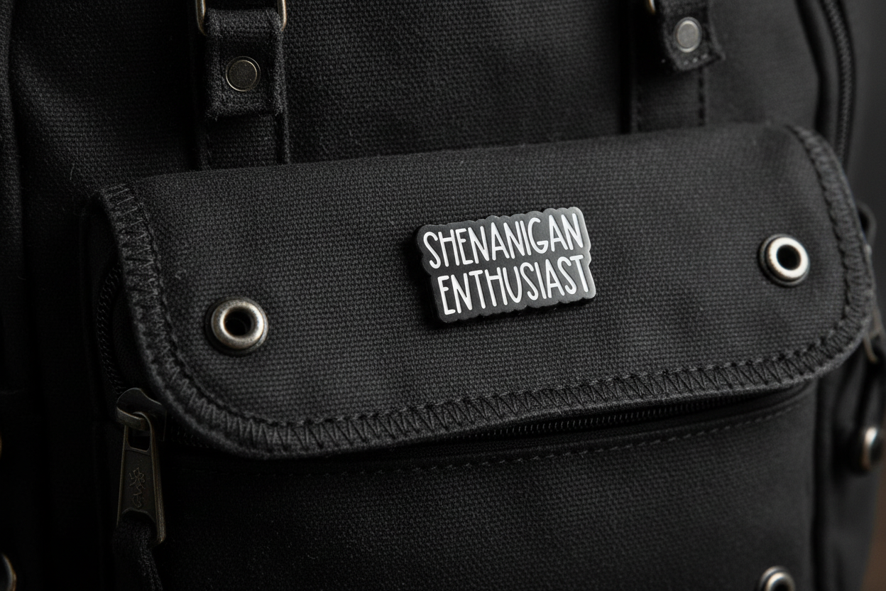 Black and white enamel pin with 'SHENANIGAN ENTHUSIAST' text on a black backpack background