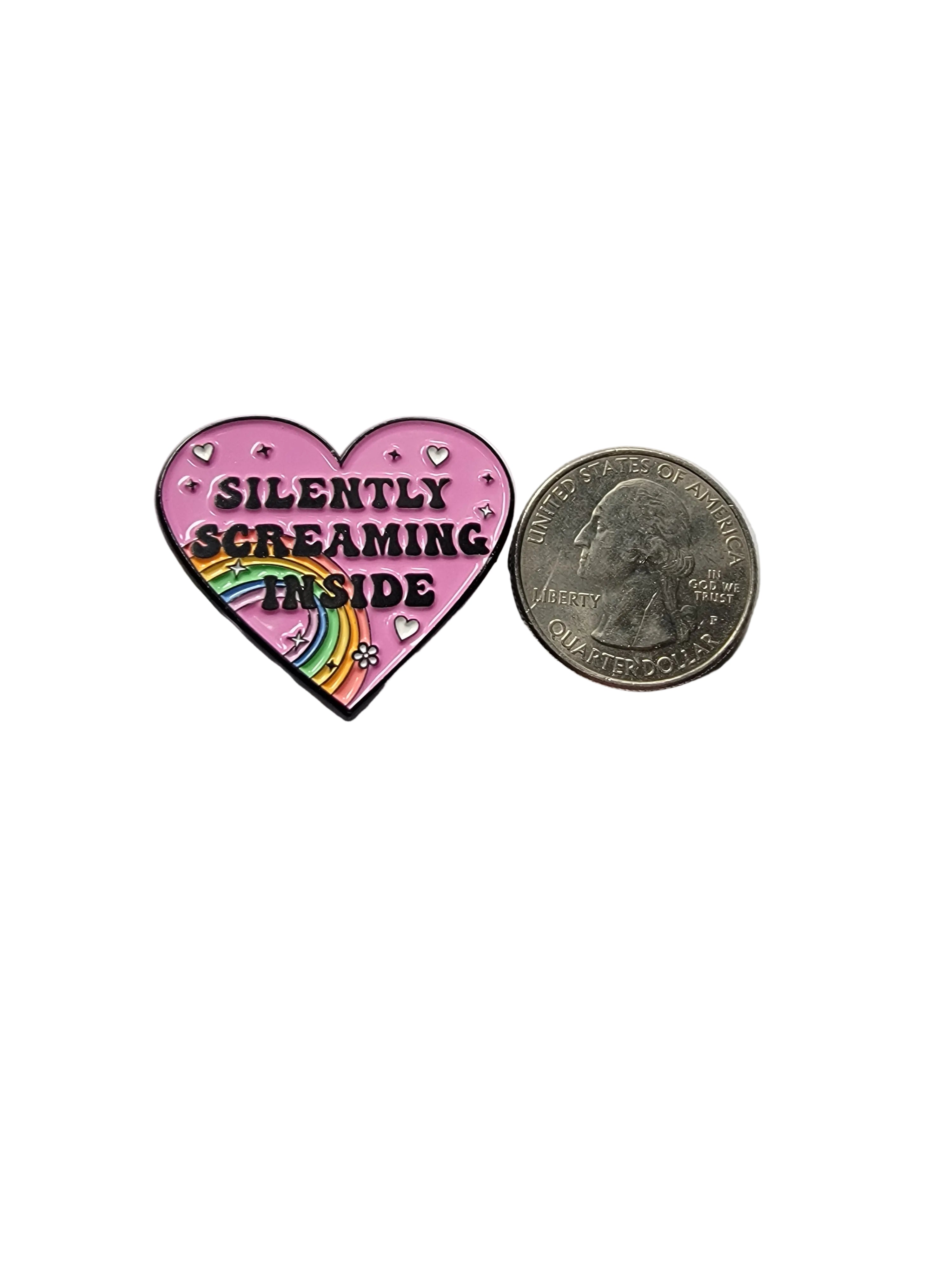 Pink heart-shaped enamel pin with rainbow and text, compared in size to a quarter on a gray background.