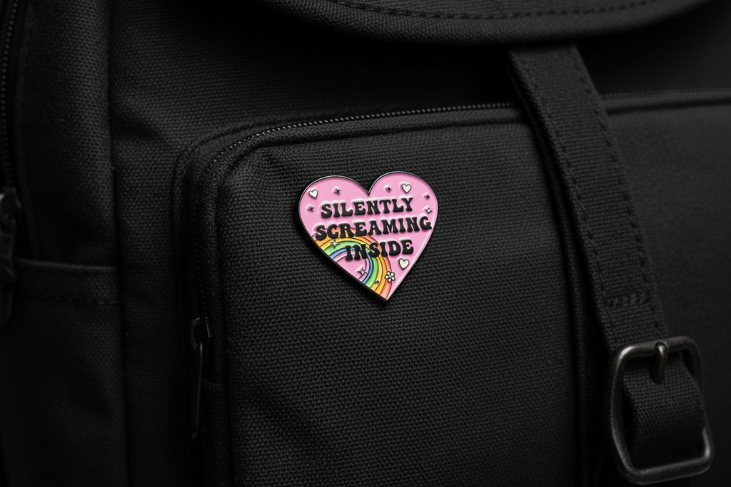Heart-shaped pink enamel pin with rainbow and text on a black bookbag background