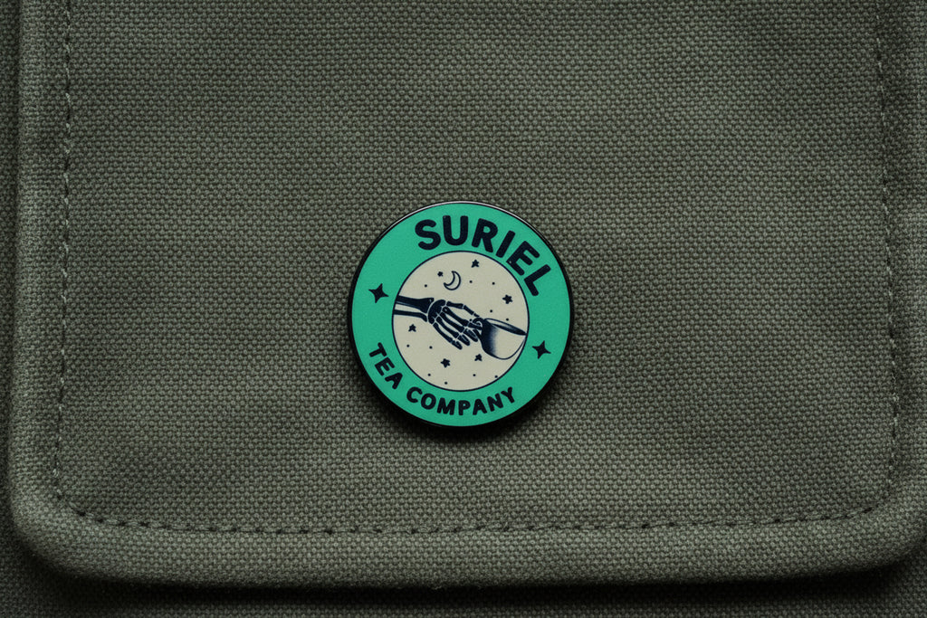 Green round pin with 'Suriel Tea Company' text on a wooden surface