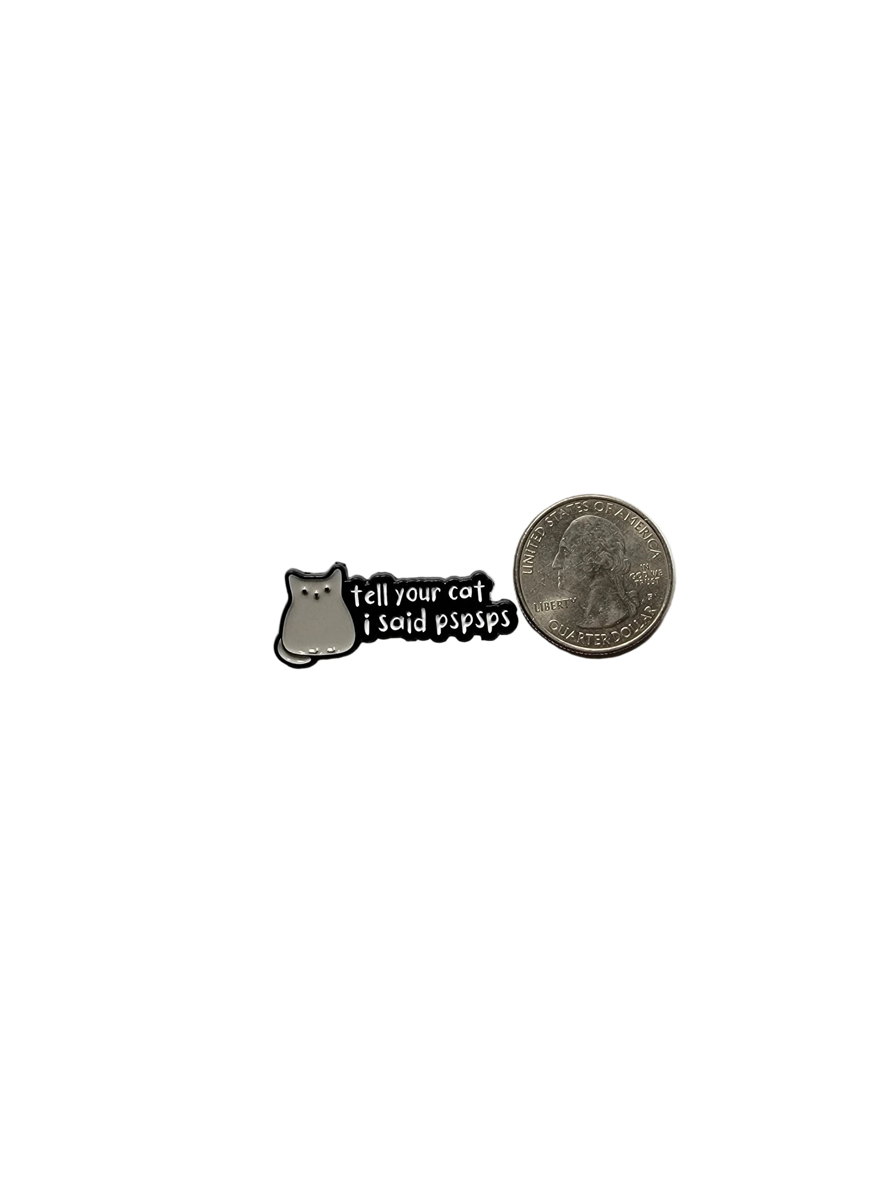 a black enamel pin with white text and cat next to a quarter on a white background