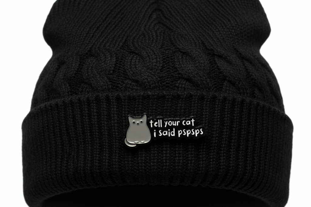 a black enamel pin with white text and a cat on a black beanie background