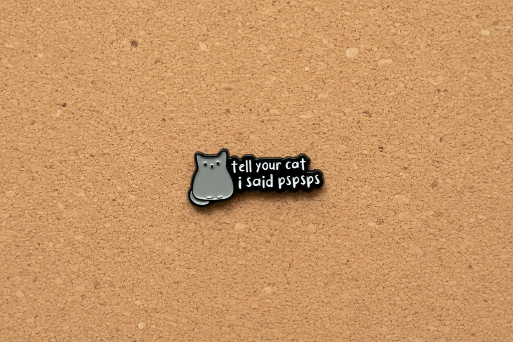 a black enamel pin with white text and a cat on a cork background