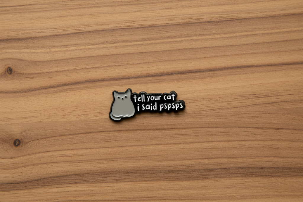 a black enamel pin with white text and a cat on a wood background