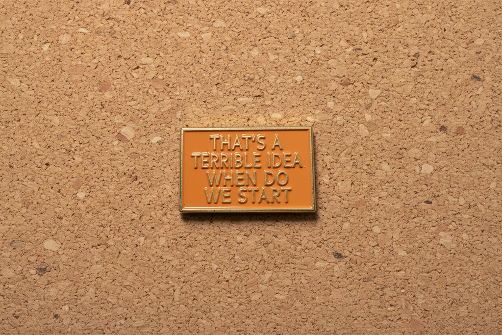 a rectangular orange pin with gold text on a cork background