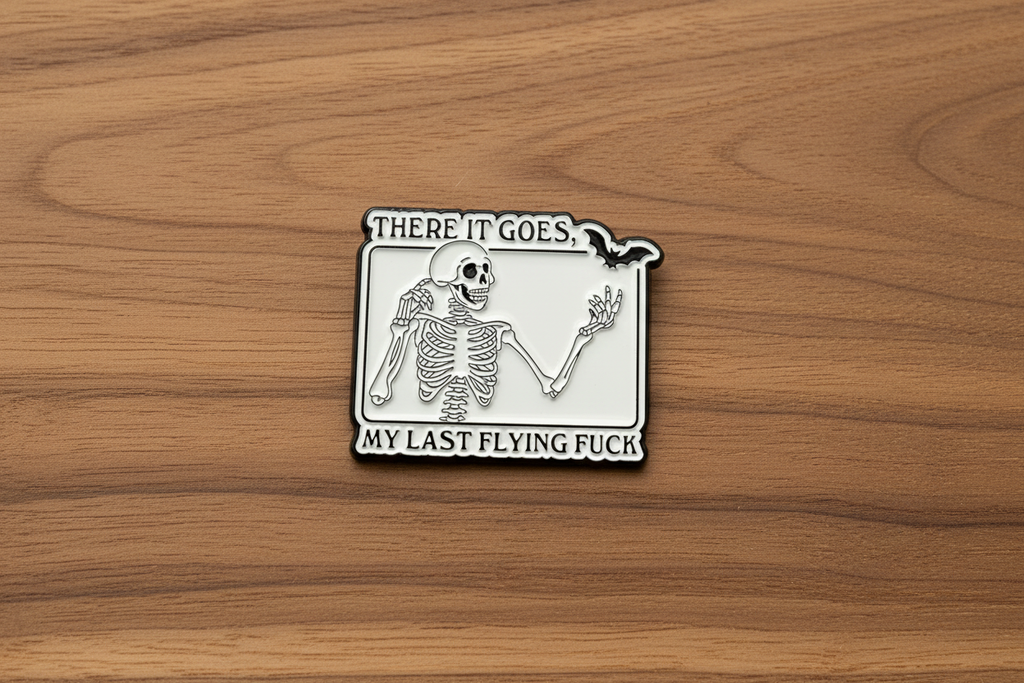 White enamel pin with skeleton and black text on a wood background. 