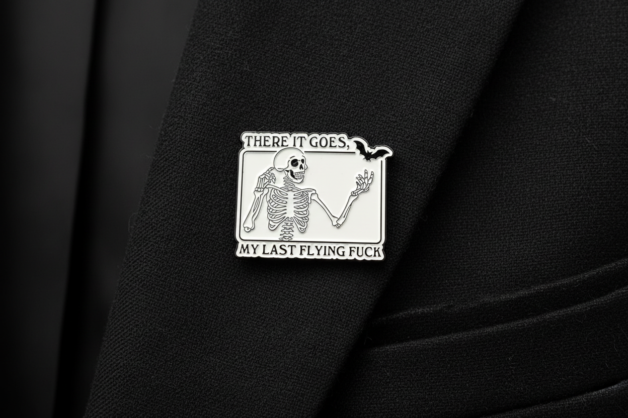 White enamel pin with skeleton and black text on a black lapel background. 