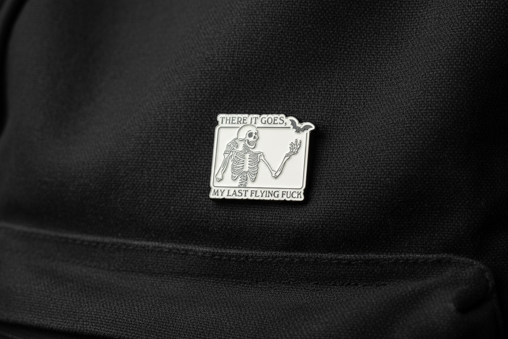 White enamel pin with skeleton and black text on a black bookbag background. 
