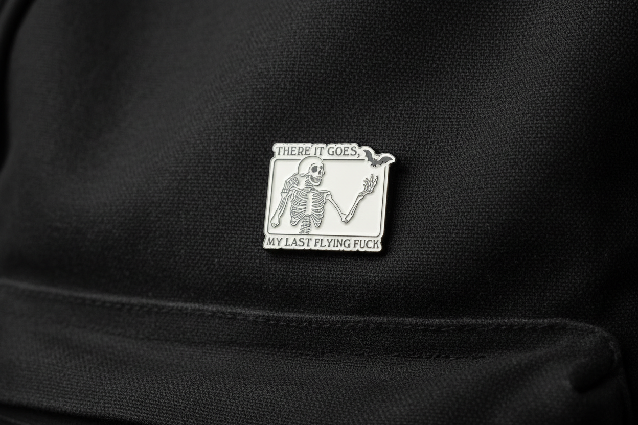 White enamel pin with skeleton and black text on a black bookbag background. 