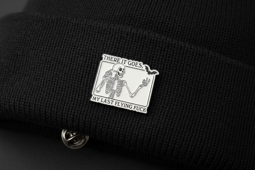 White enamel pin with skeleton and black text on a black beanie background. 