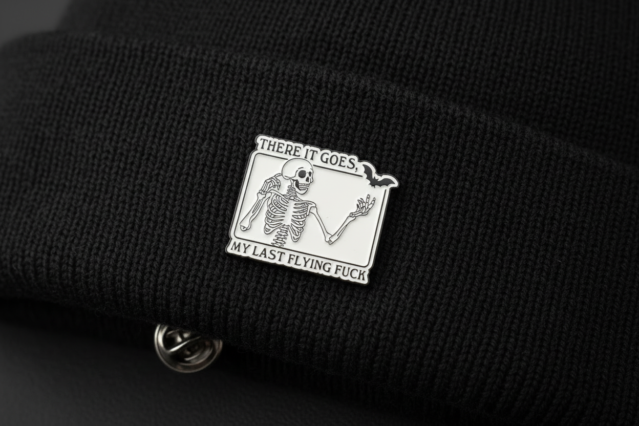 White enamel pin with skeleton and black text on a black beanie background. 