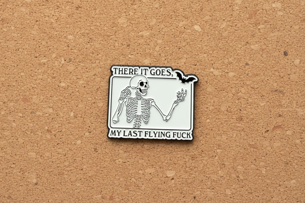 White enamel pin with skeleton and black text on a cork background. 