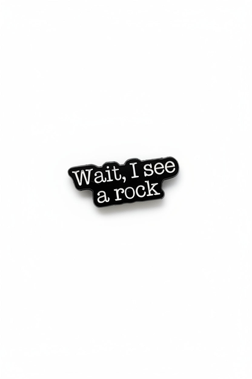 a black enamel pin with white text that says wait i see a rock on a white background